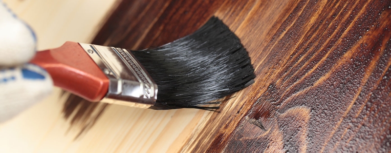 Paintbrush applying dark stain on wooden surface.